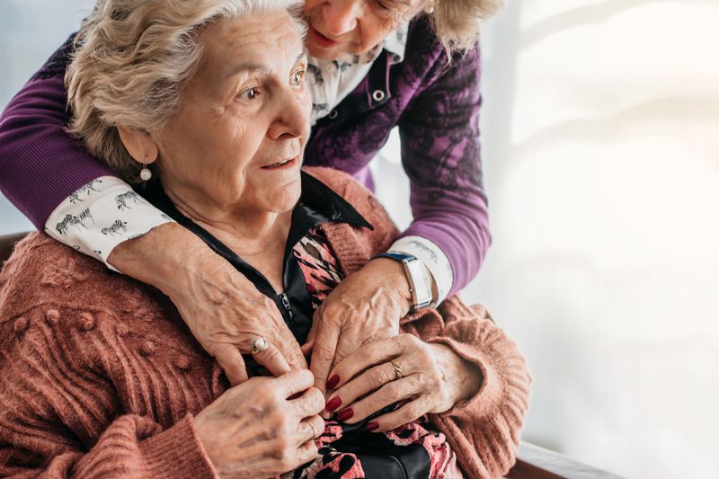 An adult daughter helps their mother adjust a sweater as they assist nursing home staff administering care.