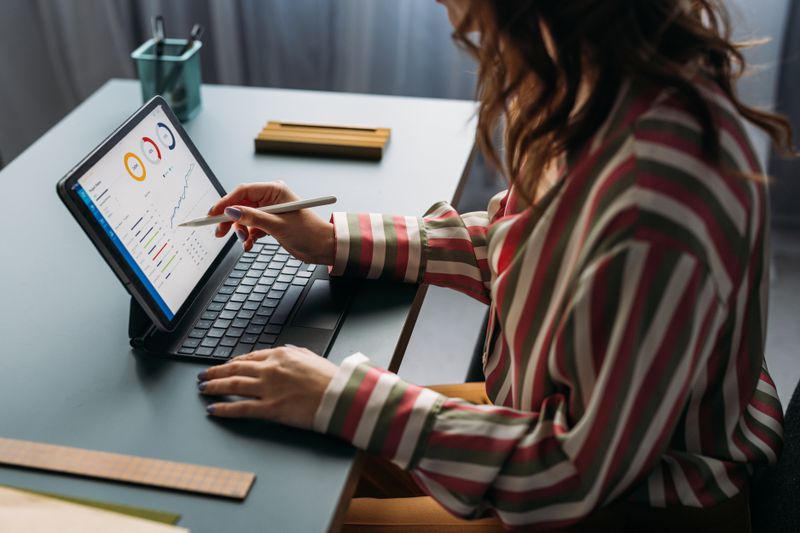 Focused young female analyzing financial statistics on digital tablet, working from a modern home office setup.
