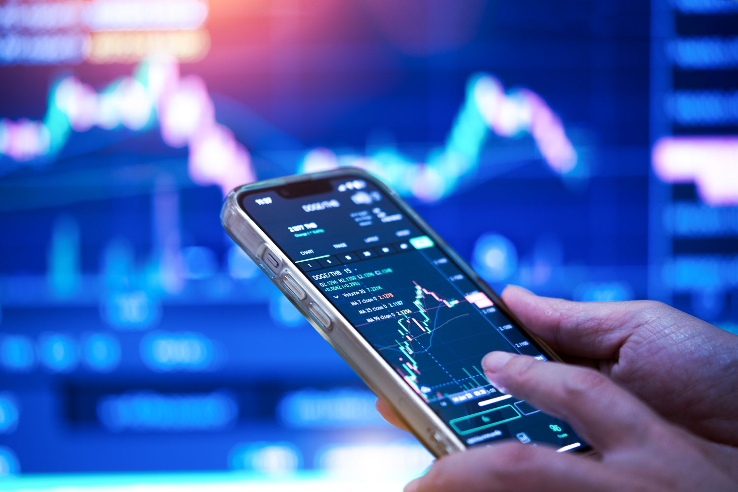 A close-up image of a person's hands holding a cell phone displaying stock market data, with a computer screen in the background.