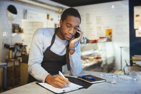 Confident young male employee talking through smart phone while writing on clipboard at delicatessen