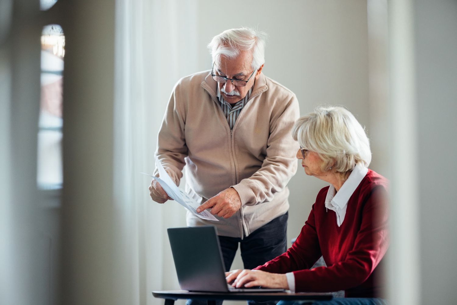 Elderly couple working together on a project while discussing documents and using a laptop in a cozy and bright environment.