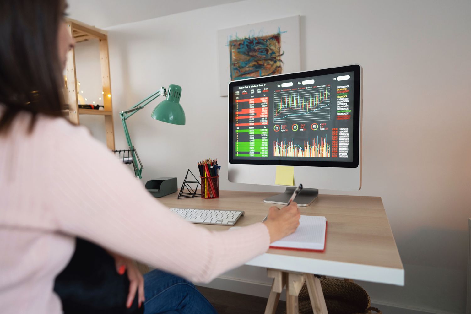 Woman looking at financial data on a monitor while writing in a note pad.