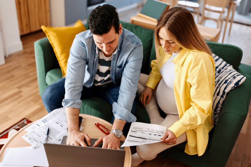 Young couple, with wife pregnant, sitting on their sofa and looking at mortgage documents and a laptop