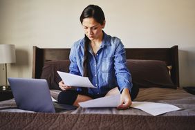 A woman sitting on her bed in a home working on a laptop.