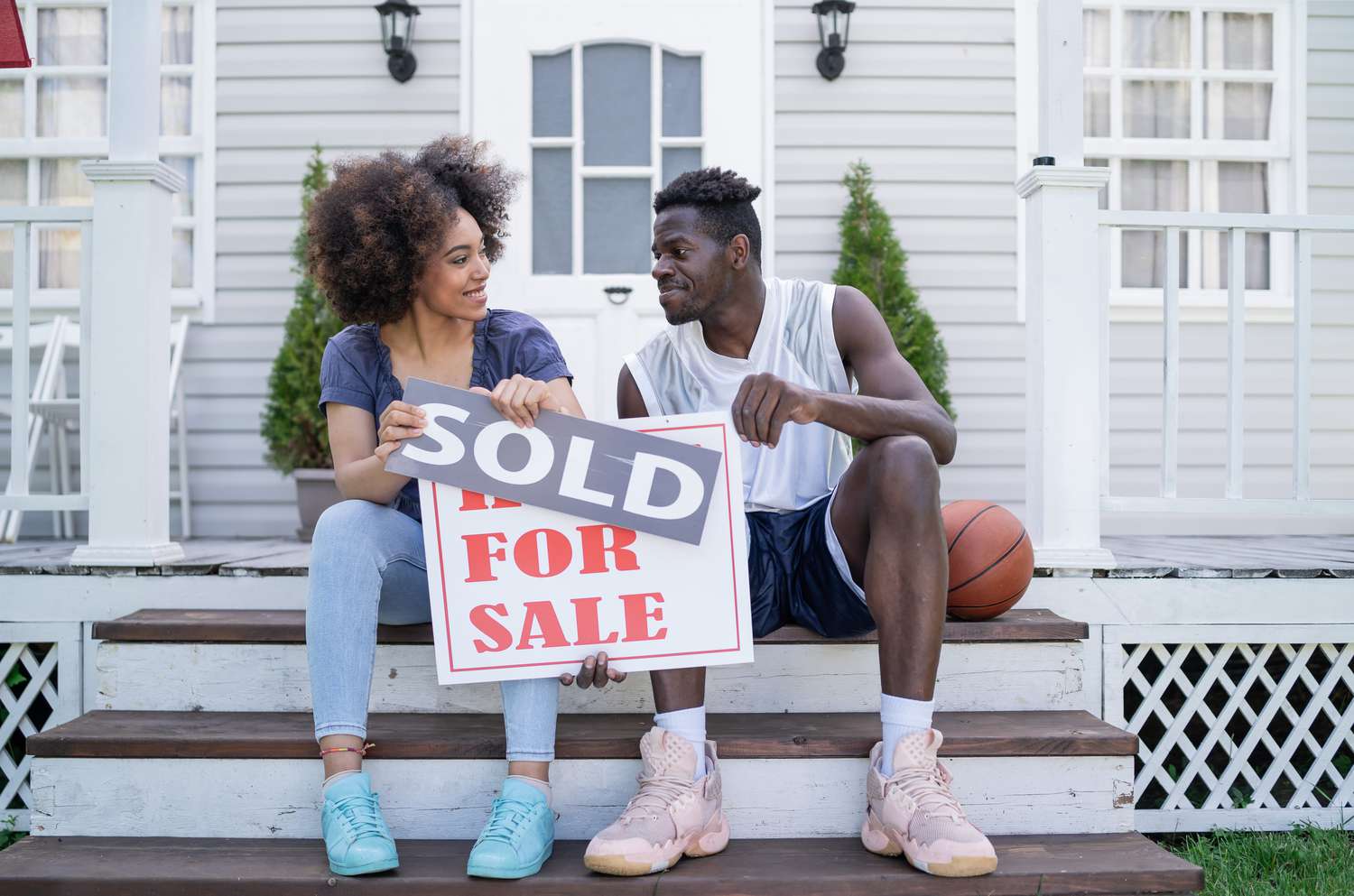 Happy couple holding "sold" signs, sitting on the porch in front of the property.