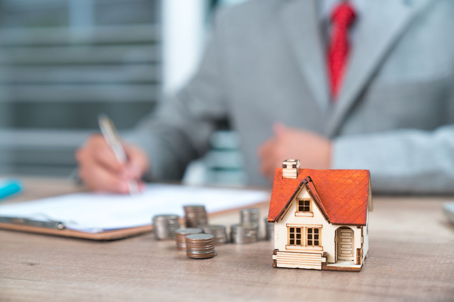 A person wearing a suit and tie signs a document at a desk, with coins and a house figurine in the foreground.