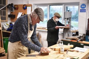 Two men work on woodworking projects in a carpentry workshop, one at a bench sanding, and the other in the background