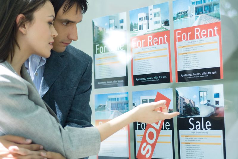 Two people looking at For Rent and For Sale property signs in a window