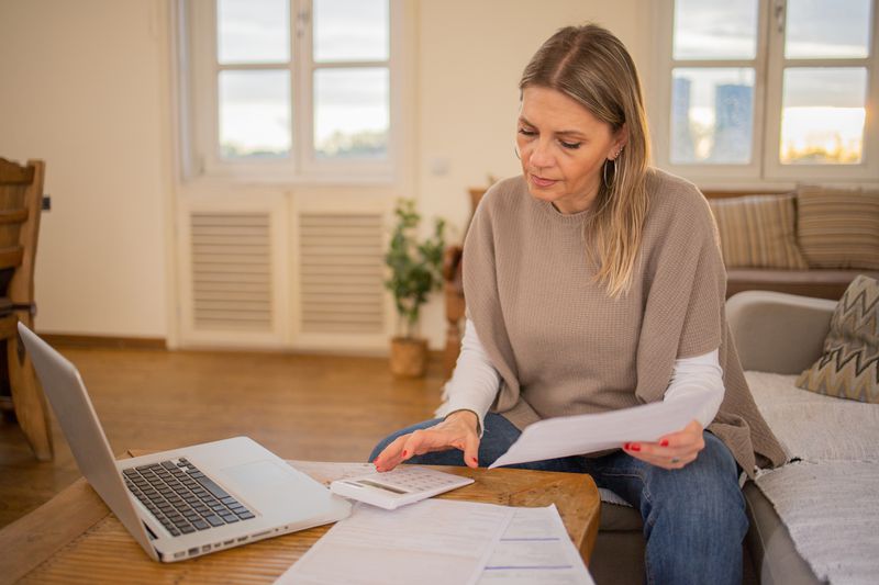 Woman in her 40s or 50s sitting on her sofa and looking at financial documents, a laptop, and a calculator