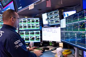 A stock trader stands at a desk looking at several computer monitors with stock charts