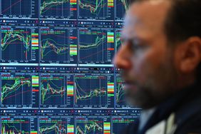 A trader's side profile is out of focus, and screens behind him display several stock charts