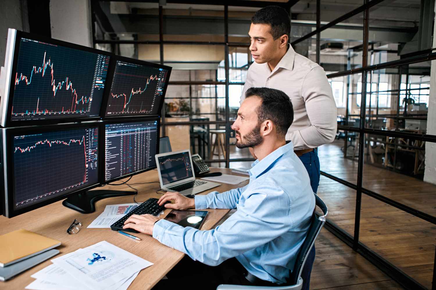 Two traders, one sitting, the other standing looking at several computer monitors displaying financial data.