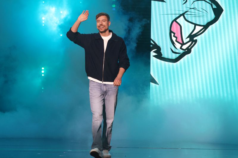 MrBeast waving while walking on stage with blue lighting and a graphic design background